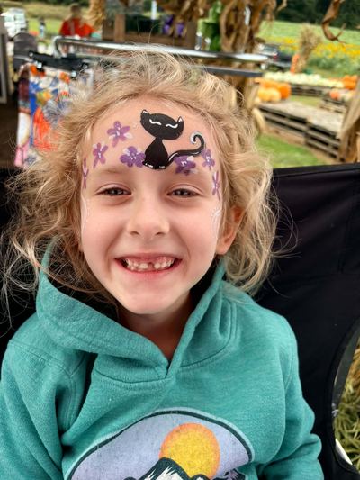 Girl with a black cat and purple flower forehead design at a fall pumpkin patch, face painting by In His Image, Morristown TN