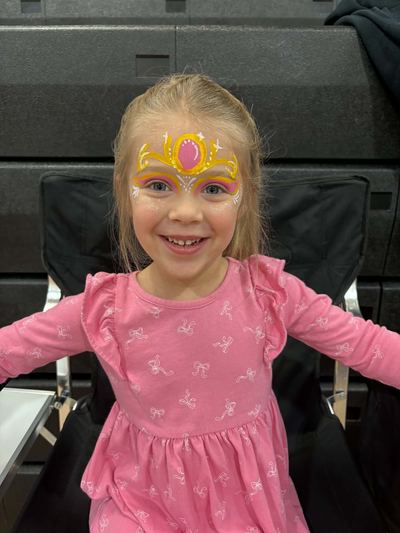 Smiling girl with a gold princess tiara and pink gem design, face painting by In His Image, Morristown TN